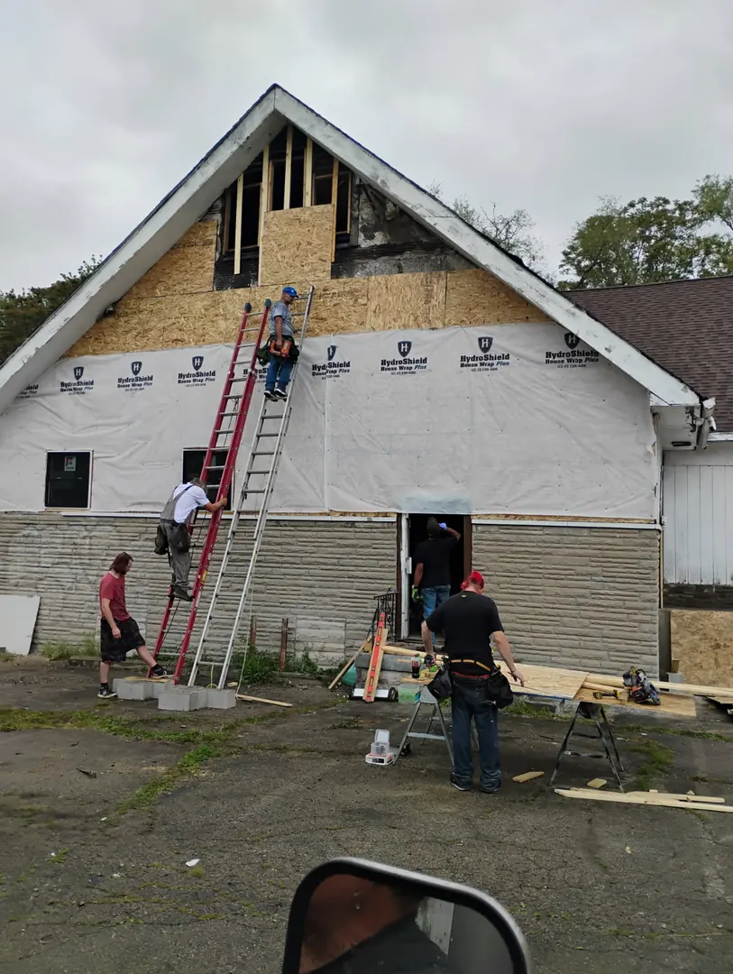Crew on ladders at a tall gable wall covered in house wrap during an exterior rebuild