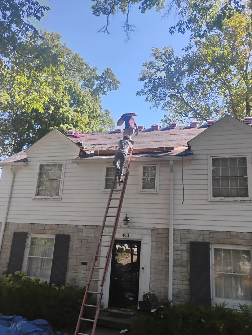 Roofer on a ladder at the eave of a two-story home during a roof tear-off