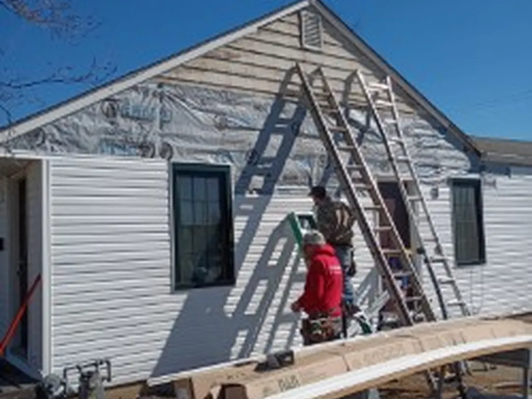 Siding installer on a ladder hanging new white vinyl siding on a gable end